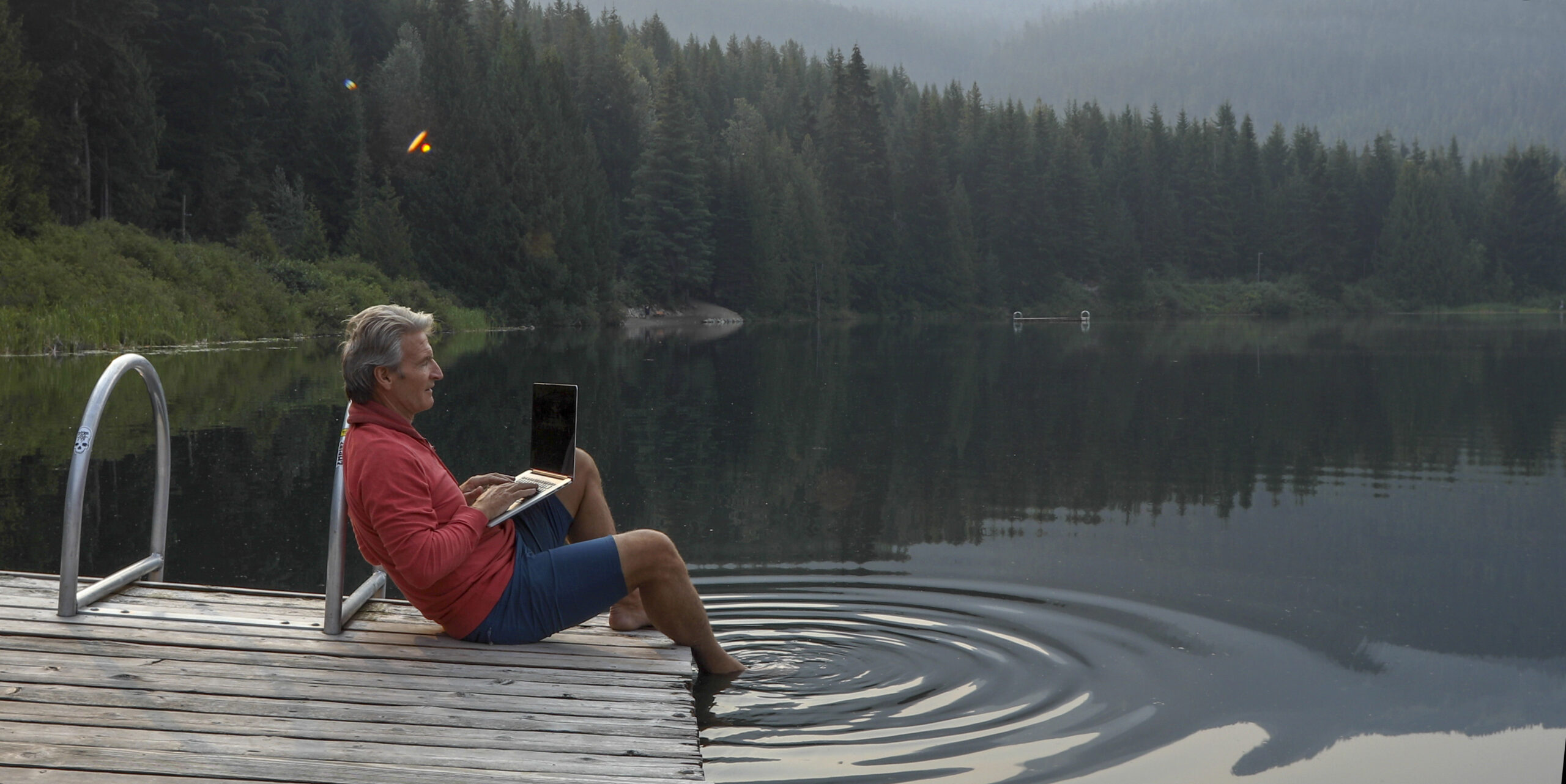 Mature man pauses on wooden pier, looks out across lake.He uses laptop computer. Lost Lake, Whistler, BC